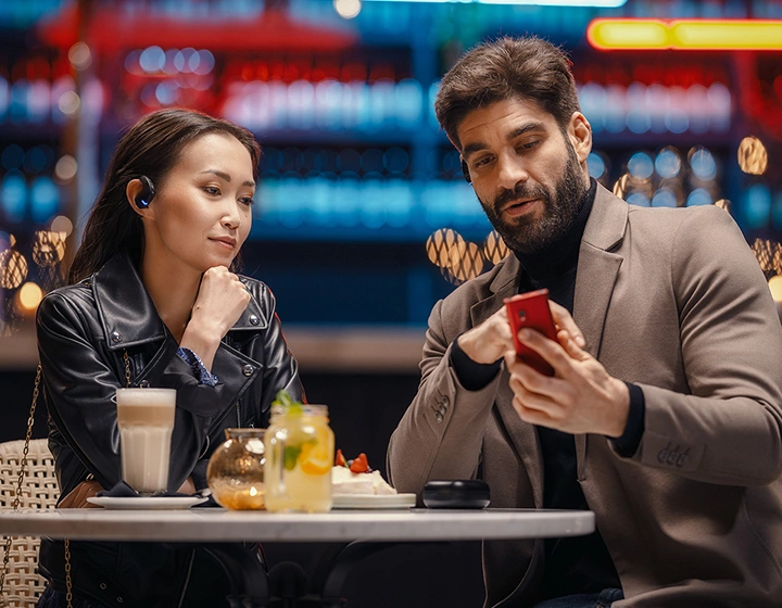 A woman and a man sitting together in a restaurant, both wearing Vasco Translator E1 earbuds; the man holds a Vasco Translator V4 and shows something to the woman.