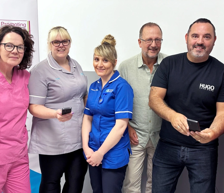 Viisi hymyilevää ihmistä seisoo yhdessä terveydenhuollon asuissa, ja kahdella heistä on tulkkilaite kädessään.