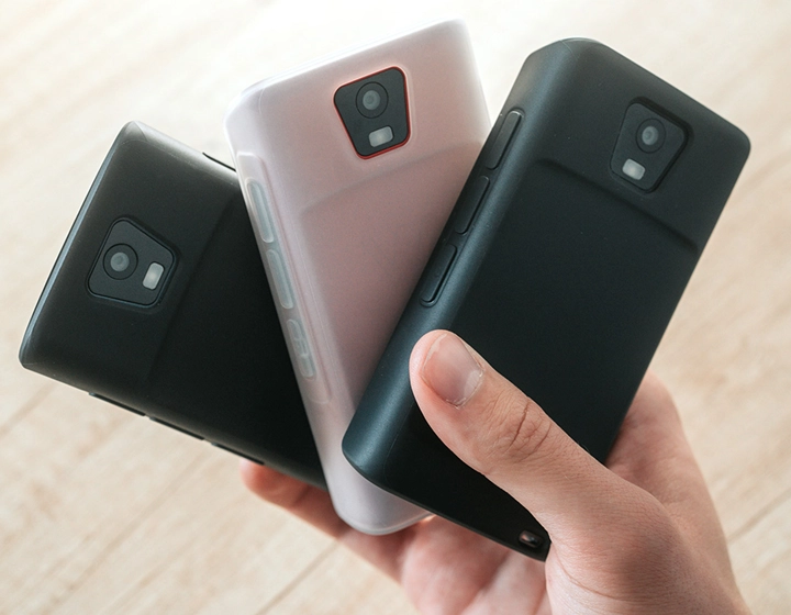 Henkilö, jolla on kädessään kolme Vasco Translator Q1 -laitetta silikonikoteloissa; kuvassa näkyy kolme eri koteloväriä: Solid Black, Dark Caramel ja Milky White.