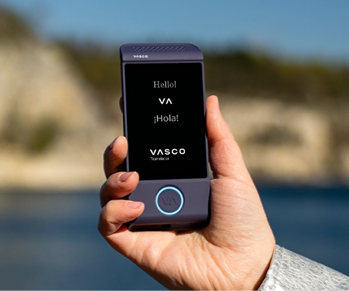 Lähikuva kädestä, joka pitelee violettia Vasco Translator M4 -käännöslaitetta kirkkaalla ulkotausalla. Näytöllä näkyy tervehdykset ”Hello!”, ”¡Hola!” sekä ”VASCO Translator” -logo.